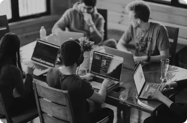 A black and white photograph of a group of young developers or students collaborating around a wooden table, working on laptops with one person wearing headphones in the foreground.
